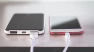 Um telemóvel cor-de-rosa e uma bateria externa preta e branca, lado a lado sobre uma mesa, ligados a cabos de carregamento brancos