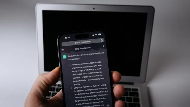 Mão a segurar um smartphone com o ChatGPT aberto no browser, à frente de um portátil prateado