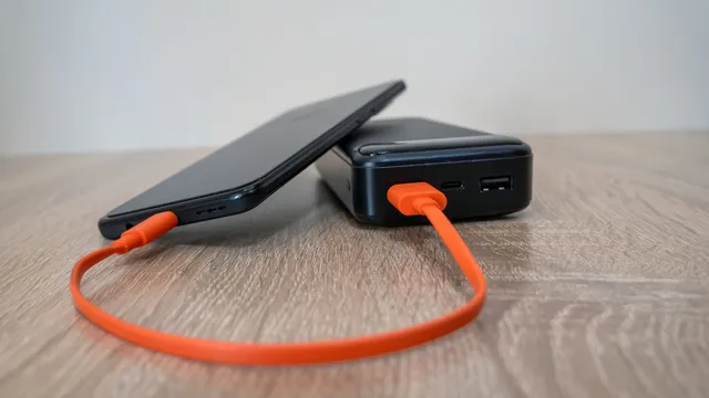 Telemóvel preto encostado a uma bateria externa preta, ligados por um cabo cor de laranja, sobre uma mesa de madeira