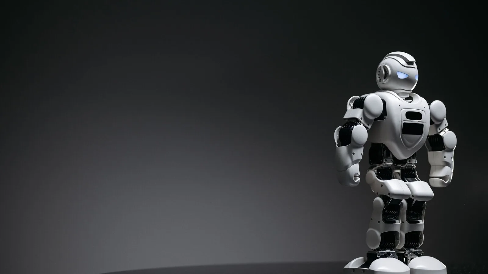 Pequeno robô de brinquedo branco e preto, de aspeto humanoide e olhos azuis acesos, sobre um fundo cinzento escuro