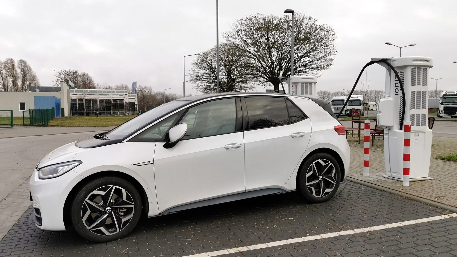 Volkswagen ID.3 branco estacionado junto a um posto de carregamento rápido, num parque de estacionamento com árvores sem folhas