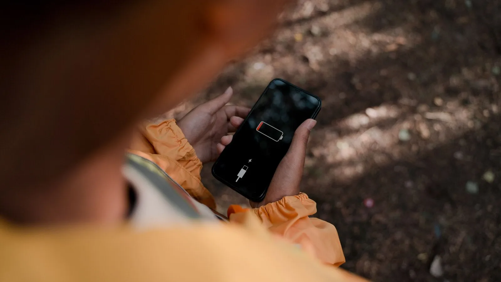 Pessoa de casaco cor de laranja segura um telemóvel com o ícone de bateria vazia no ecrã, sobre um fundo de terra