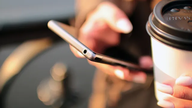 Mão a segurar um telemóvel prateado, vista de perfil, ao lado de um copo de café para levar