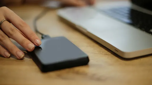 Mão pousada sobre um disco externo preto ligado por cabo a um portátil prateado, numa mesa de madeira