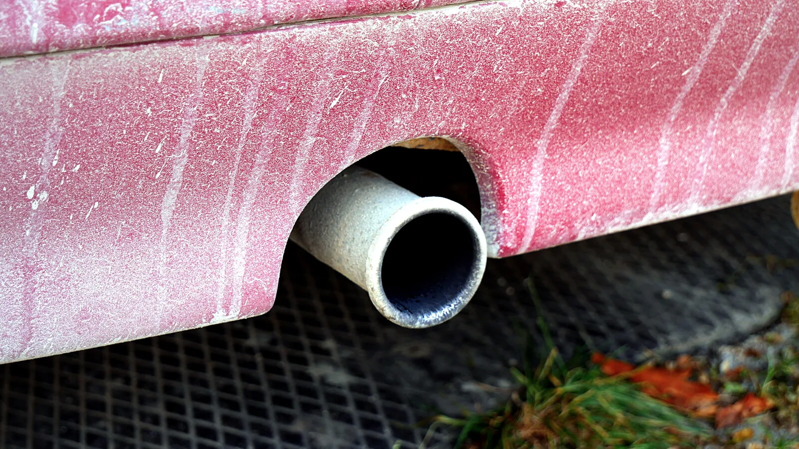 Tubo de escape de um carro vermelho coberto de pó, visto de perto, por cima de uma grelha metálica com folhas e erva