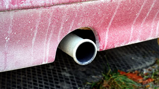Tubo de escape de um carro vermelho coberto de pó, visto de perto, por cima de uma grelha metálica com folhas e erva
