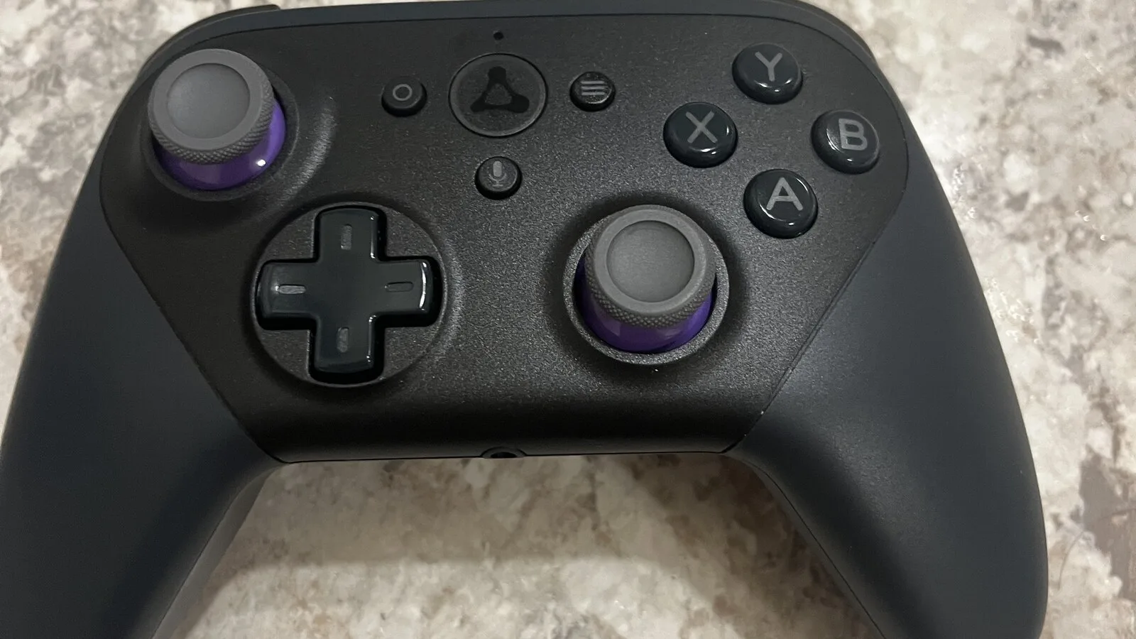 Comando de jogos preto com manípulos de anel roxo e o símbolo triangular da Luna ao centro, pousado numa bancada de pedra