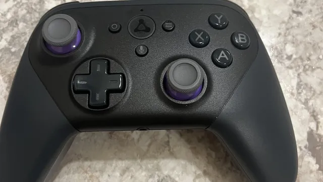 Comando de jogos preto com manípulos de anel roxo e o símbolo triangular da Luna ao centro, pousado numa bancada de pedra