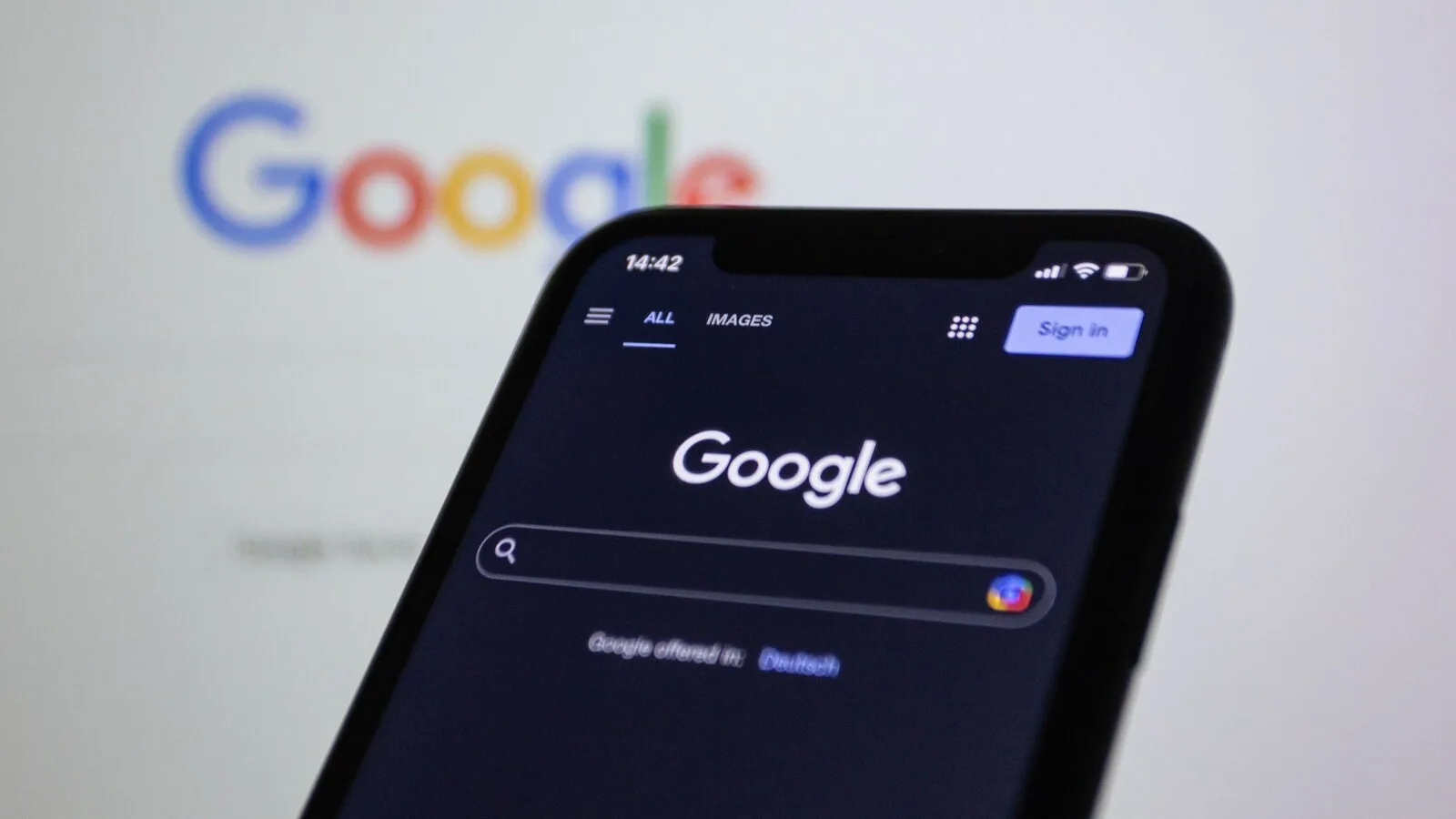 Telemóvel com a página inicial da Pesquisa Google em modo escuro, à frente de um ecrã desfocado com o logótipo da Google