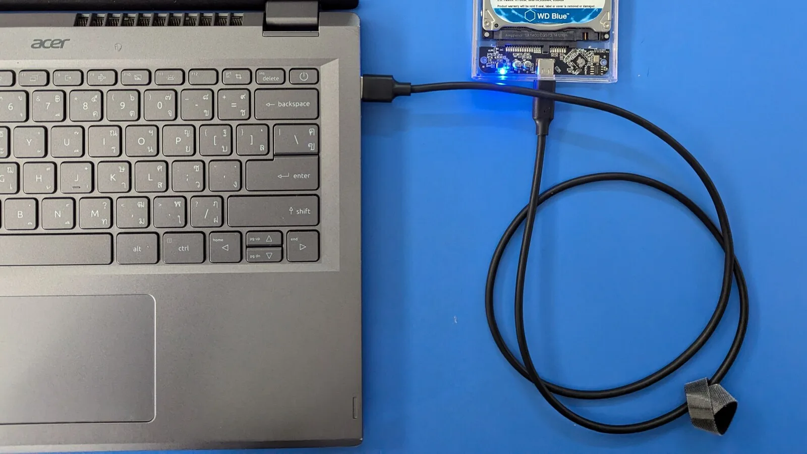 Disco rígido externo numa caixa transparente ligado por cabo USB-C a um portátil cinzento, sobre uma mesa azul