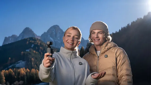 Uma mulher segura uma câmara DJI Osmo Pocket 4 à frente do rosto e filma-se ao lado de um homem de gorro, com montanhas e um pinhal ao fundo