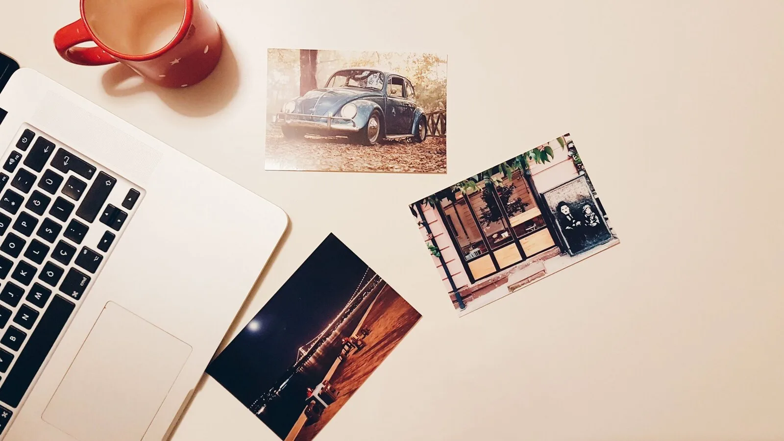 Portátil prateado aberto numa secretária branca, ao lado de uma caneca vermelha e de três fotografias impressas