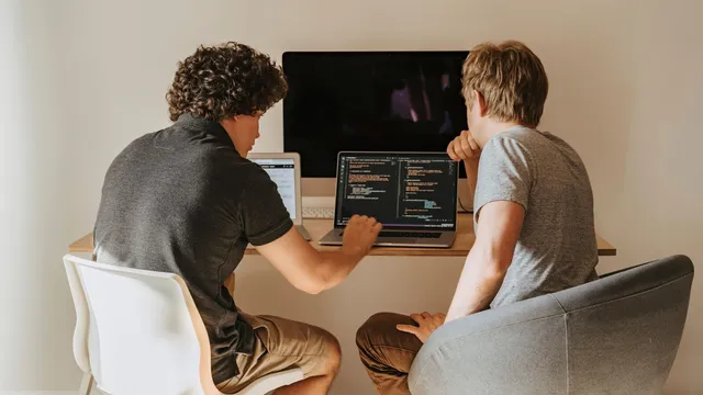 Dois jovens de costas sentados a uma secretária, a olhar para código num portátil, com um monitor e um tablet ao lado