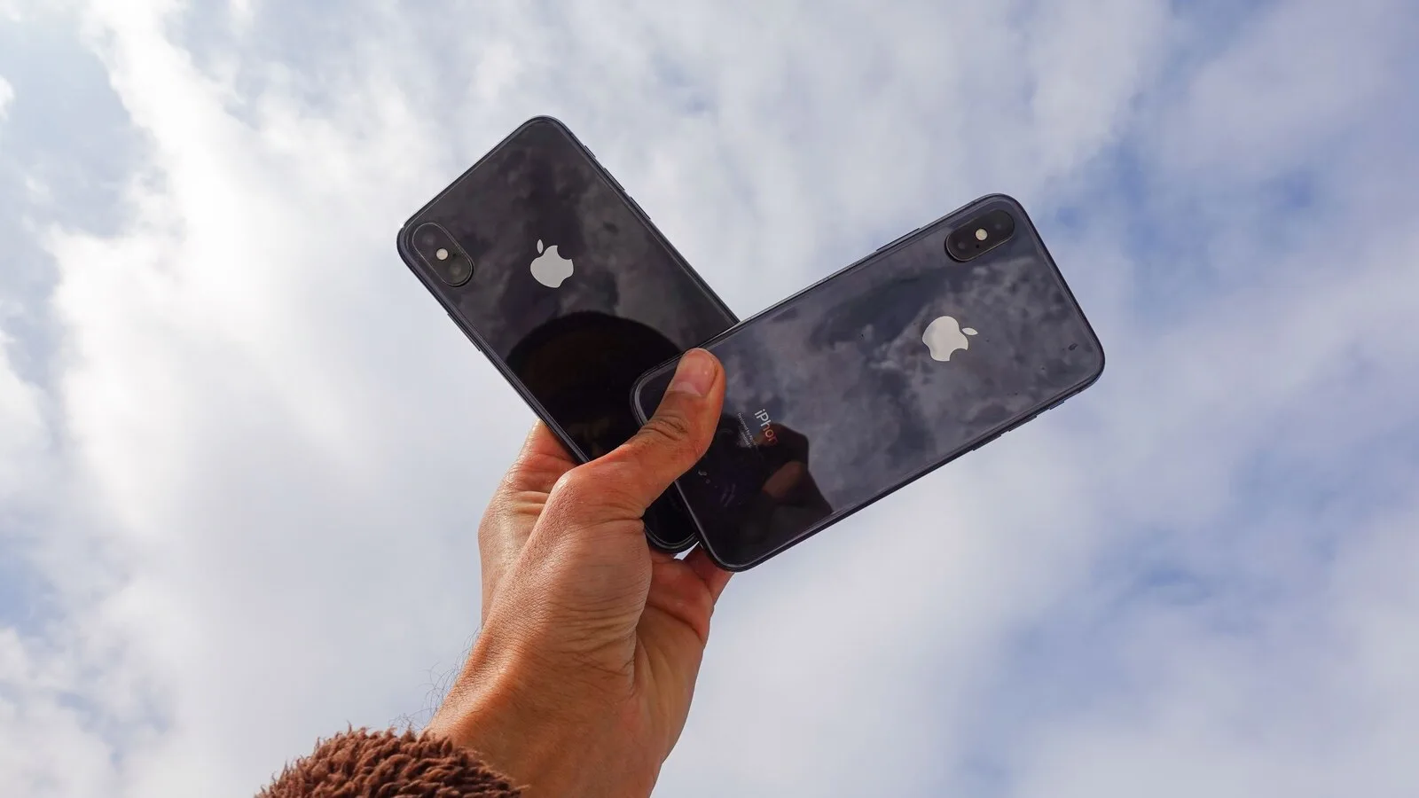 Mão a segurar dois iPhone pretos, vistos de trás e sobrepostos, contra um céu azul com nuvens