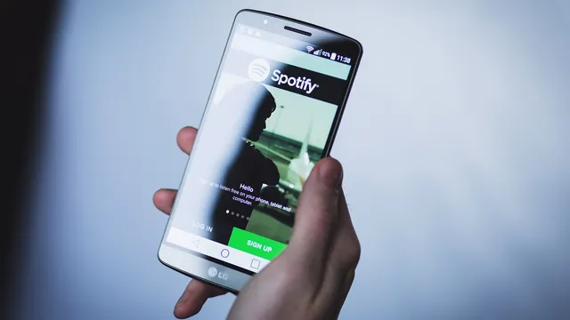 Mão a segurar um telemóvel com a app do Spotify aberta no ecrã de entrada, com fundo azulado desfocado