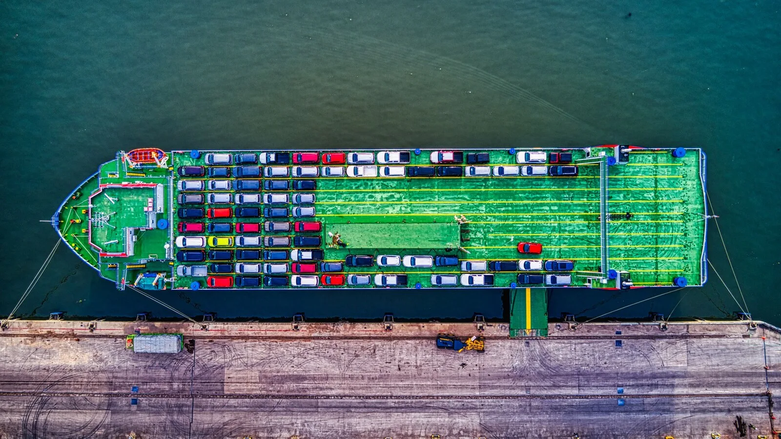Vista aérea de um navio de transporte de automóveis atracado num cais, com o convés verde cheio de carros de várias cores alinhados