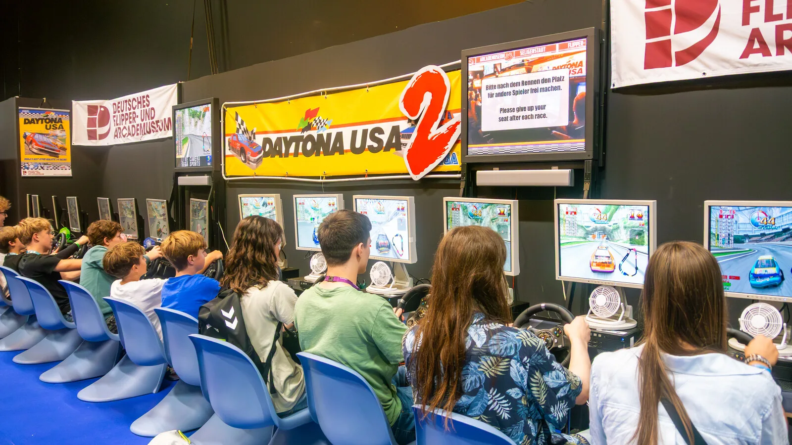 Fila de jovens sentados em cadeiras azuis a jogar em máquinas de corridas com volantes, numa feira de videojogos