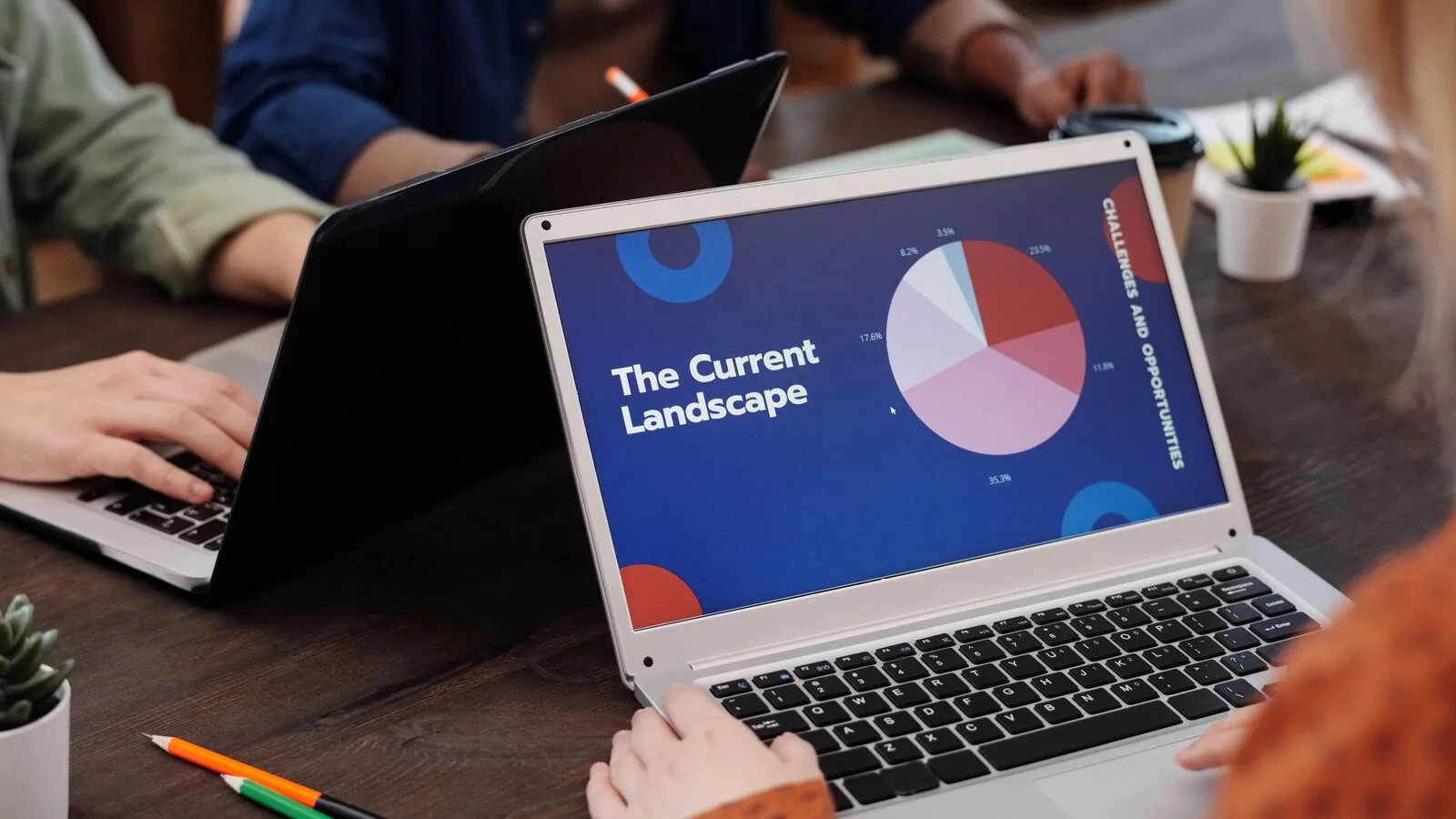 Portátil prateado numa mesa de madeira a mostrar um diapositivo azul com um gráfico circular, com outras pessoas a trabalhar em portáteis ao lado