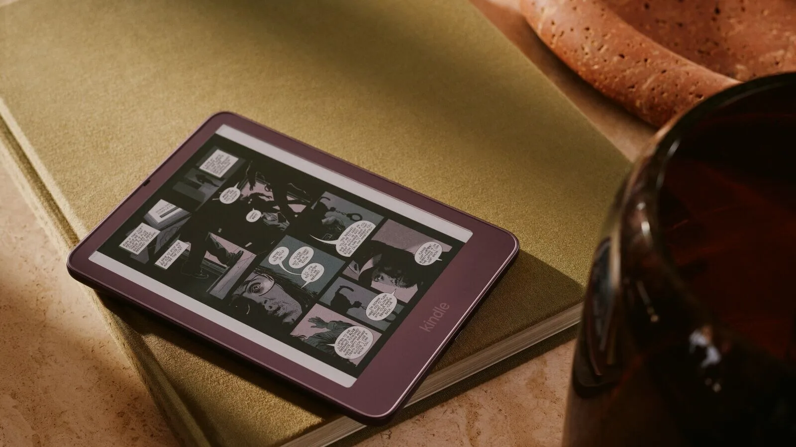 Um Kindle cor de vinho pousado sobre um livro de capa verde, com uma banda desenhada a preto e branco no ecrã, ao lado de um copo de vidro escuro