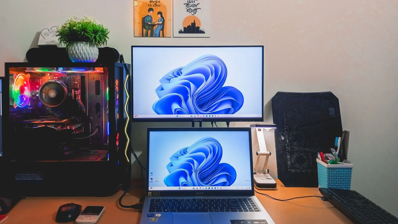 Secretária com um monitor e um portátil empilhados, ambos com o fundo azul do Windows 11, ao lado de um computador de torre com luzes coloridas