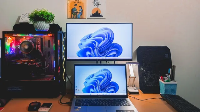 Secretária com um monitor e um portátil empilhados, ambos com o fundo azul do Windows 11, ao lado de um computador de torre com luzes coloridas