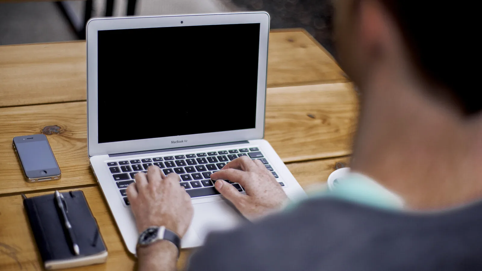 Homem a escrever num portátil MacBook prateado pousado numa mesa de madeira, ao lado de um caderno com caneta e de um telemóvel