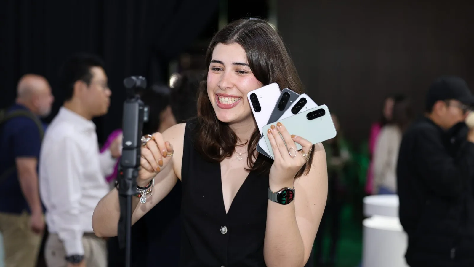 Uma jovem sorridente segura em leque quatro telemóveis Samsung Galaxy da série A, em cores claras, diante de uma câmara num tripé, num evento com público ao fundo