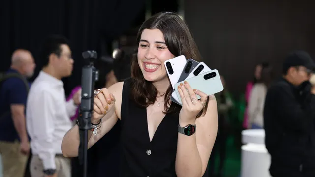 Uma jovem sorridente segura em leque quatro telemóveis Samsung Galaxy da série A, em cores claras, diante de uma câmara num tripé, num evento com público ao fundo