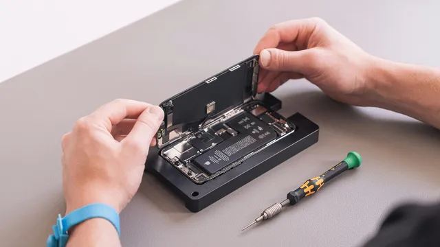 Duas mãos levantam o ecrã de um iPhone aberto, preso num suporte de reparação preto, com a bateria à vista e uma chave de parafusos pousada ao lado numa mesa cinzenta
