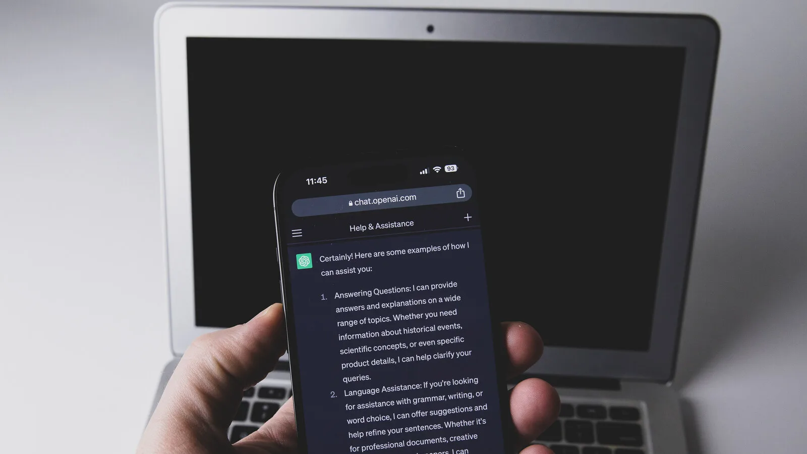 Mão a segurar um telemóvel com uma conversa do ChatGPT aberta no browser, em frente a um portátil com o ecrã apagado