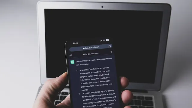 Mão a segurar um telemóvel com uma conversa do ChatGPT aberta no browser, em frente a um portátil com o ecrã apagado