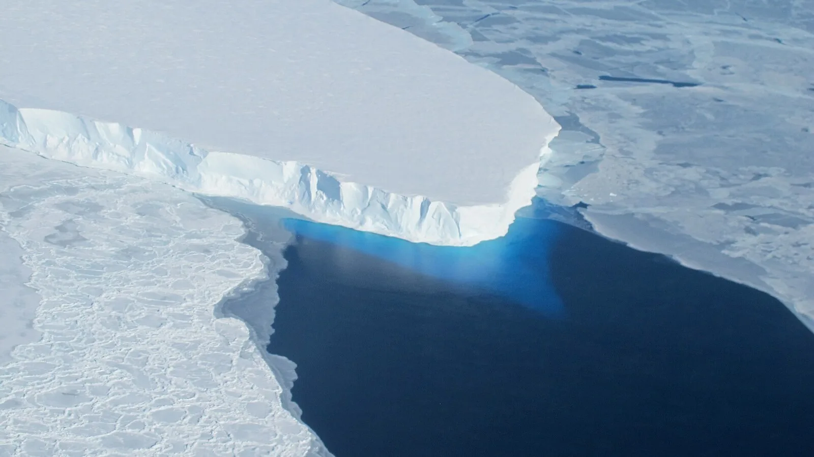 Vista aérea da frente do glaciar Thwaites, uma parede de gelo branco a terminar num mar escuro coberto de placas de gelo partido