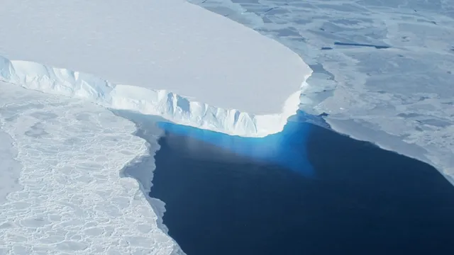 Vista aérea da frente do glaciar Thwaites, uma parede de gelo branco a terminar num mar escuro coberto de placas de gelo partido