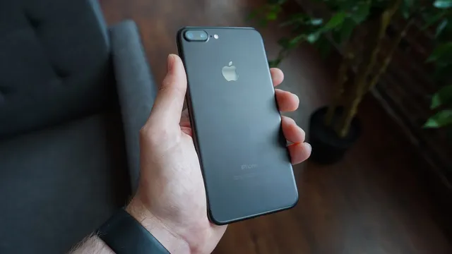 Mão de um homem com relógio no pulso a segurar um iPhone preto visto de costas, numa sala com sofá e plantas desfocados