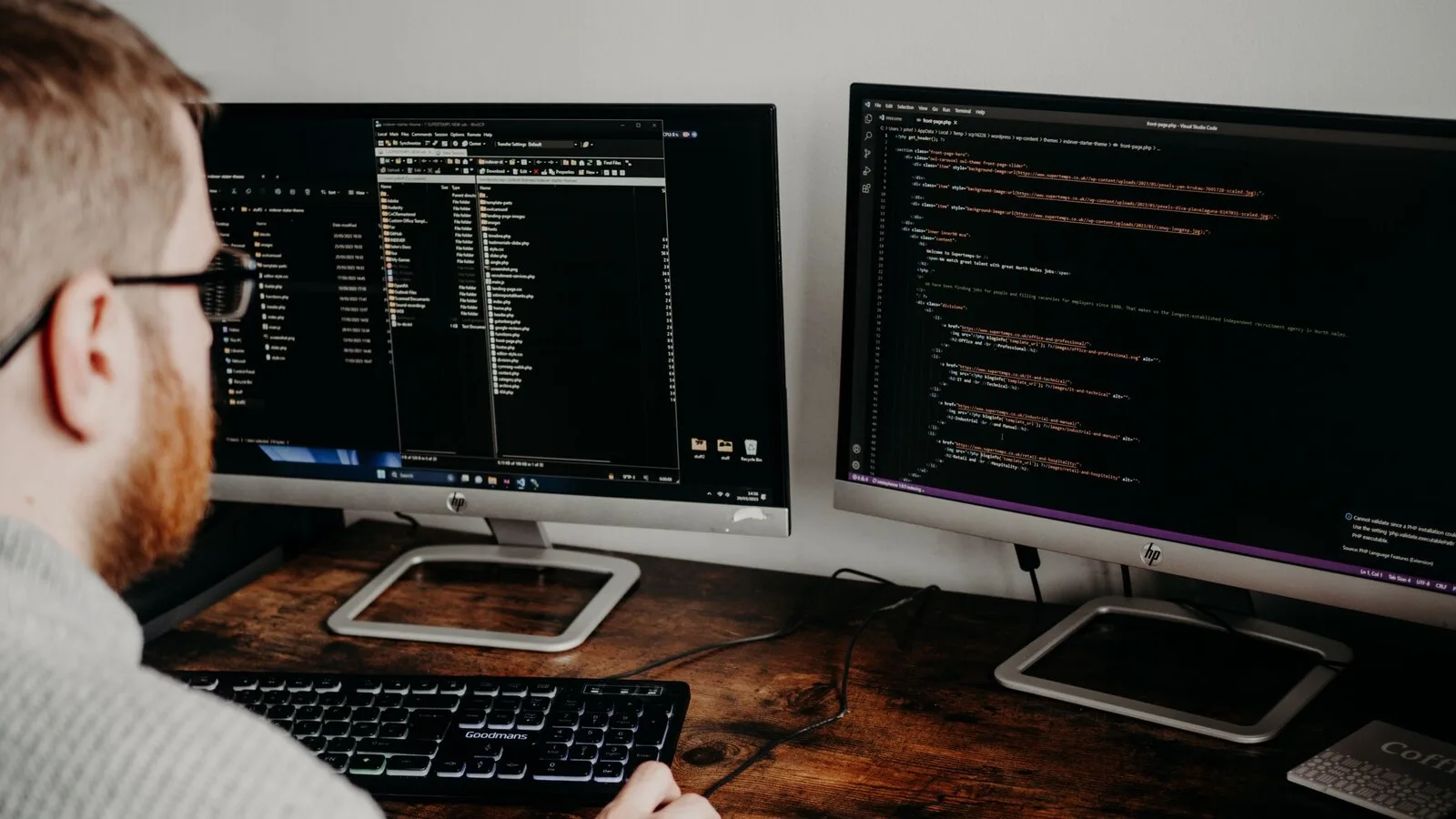 Programador de óculos sentado a uma secretária de madeira, de costas, a olhar para dois monitores com código e pastas de ficheiros