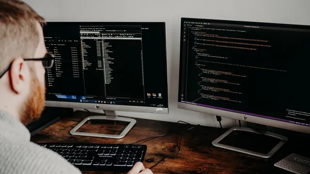 Programador de óculos sentado a uma secretária de madeira, de costas, a olhar para dois monitores com código e pastas de ficheiros