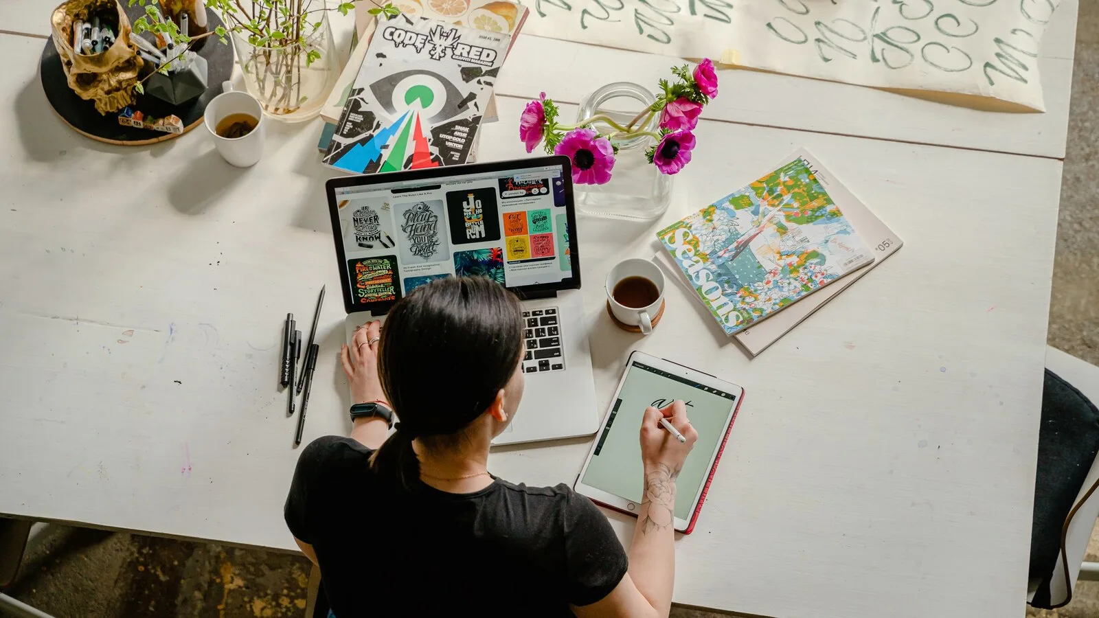 Mulher vista de cima a trabalhar numa mesa branca com um portátil aberto em páginas de tipografia, um tablet onde escreve com uma caneta, revistas, flores e chávenas de café