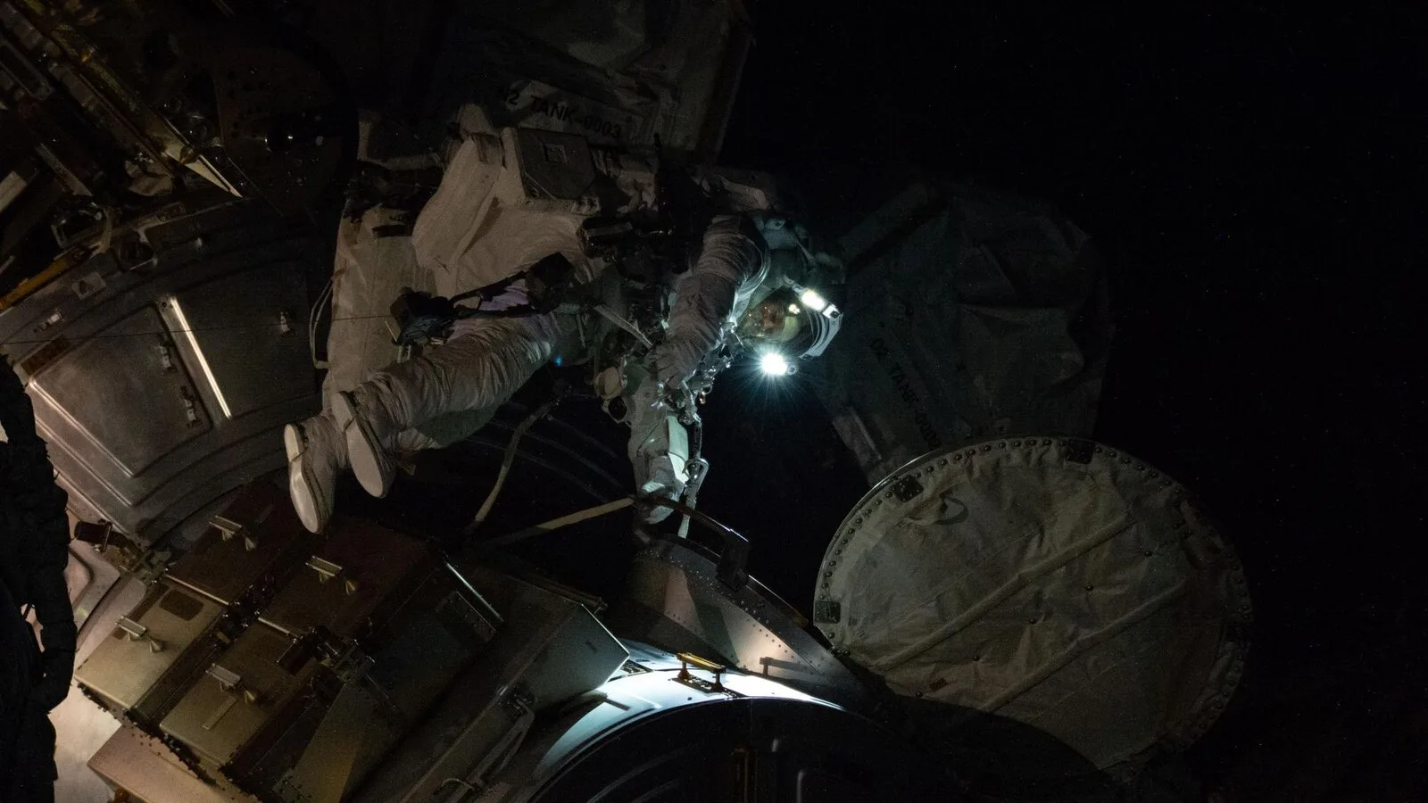 Astronauta de fato branco com as luzes do capacete acesas, no exterior da Estação Espacial Internacional, rodeado de estruturas metálicas e da escuridão do espaço