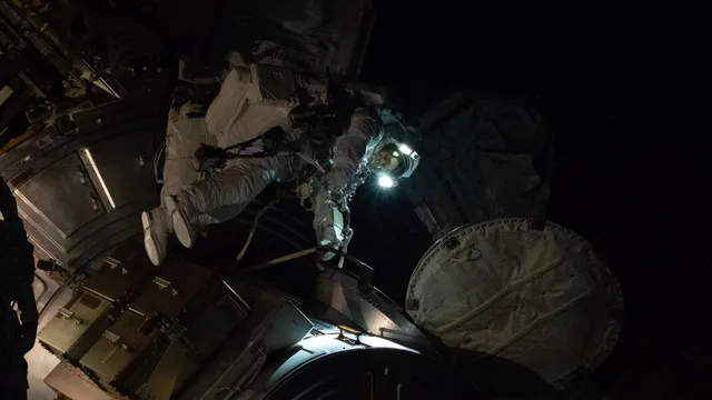 Astronauta de fato branco com as luzes do capacete acesas, no exterior da Estação Espacial Internacional, rodeado de estruturas metálicas e da escuridão do espaço