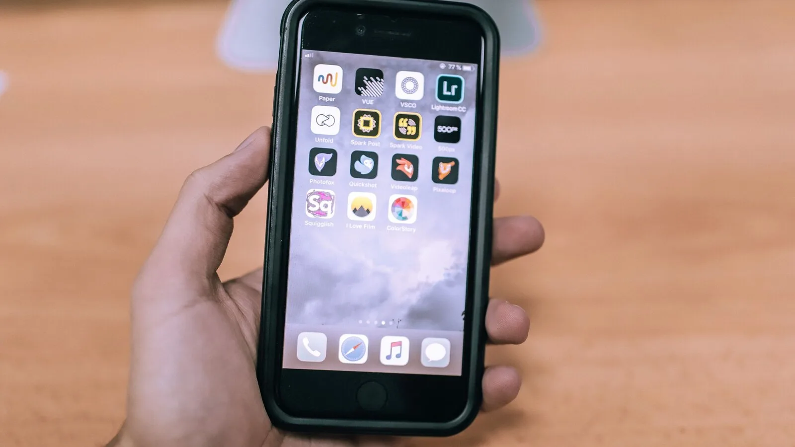 Mão a segurar um iPhone com capa preta e o ecrã inicial cheio de ícones de aplicações, sobre uma mesa de madeira desfocada