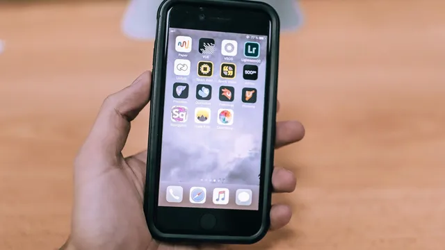 Mão a segurar um iPhone com capa preta e o ecrã inicial cheio de ícones de aplicações, sobre uma mesa de madeira desfocada