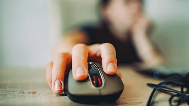 Mão pousada sobre um rato de computador escuro com luz vermelha, numa mesa de madeira, com uma pessoa desfocada ao fundo