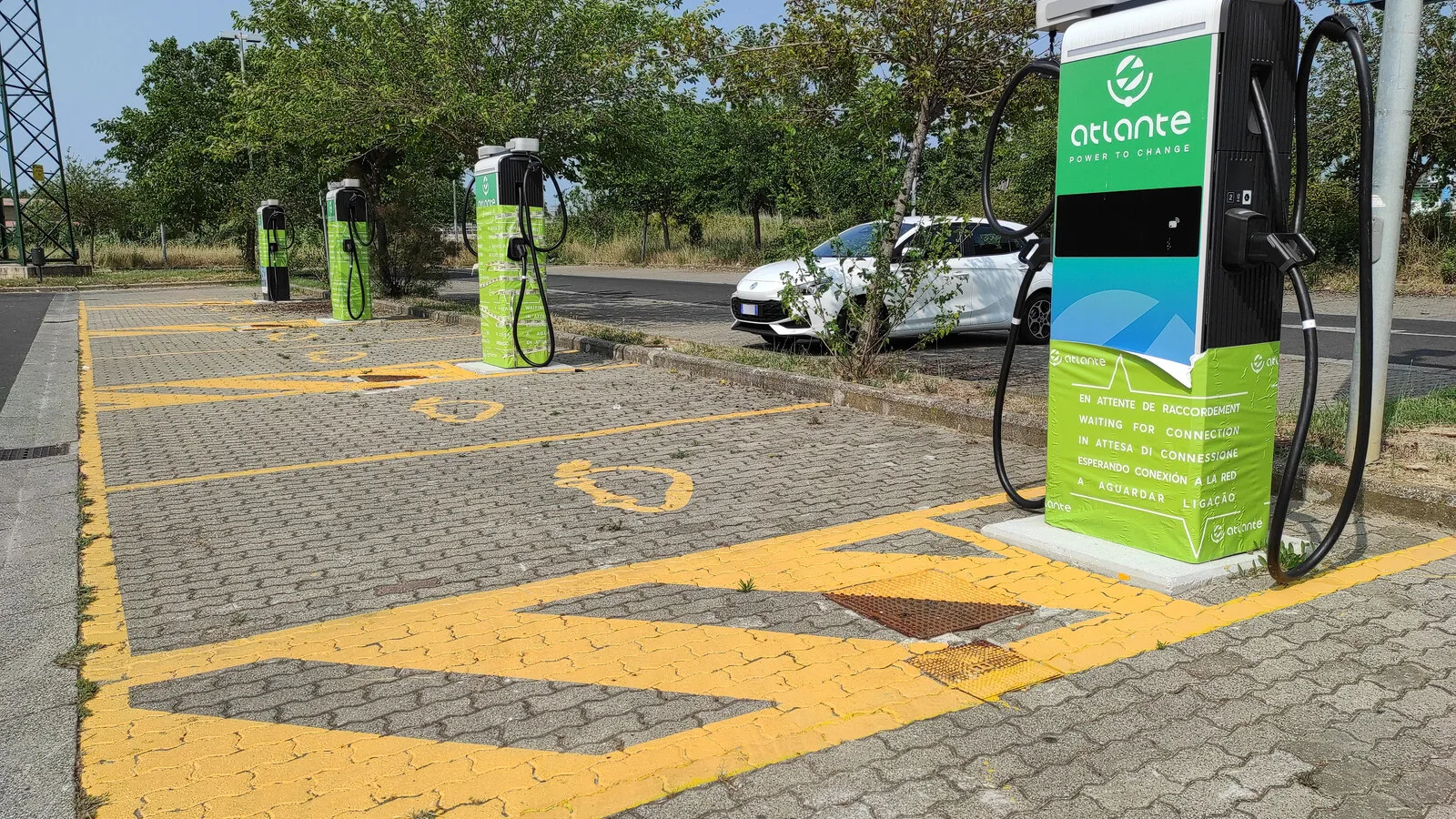 Fila de quatro postos de carregamento rápido verdes e brancos da Atlante num parque de estacionamento com lugares pintados de amarelo e um carro branco ao fundo