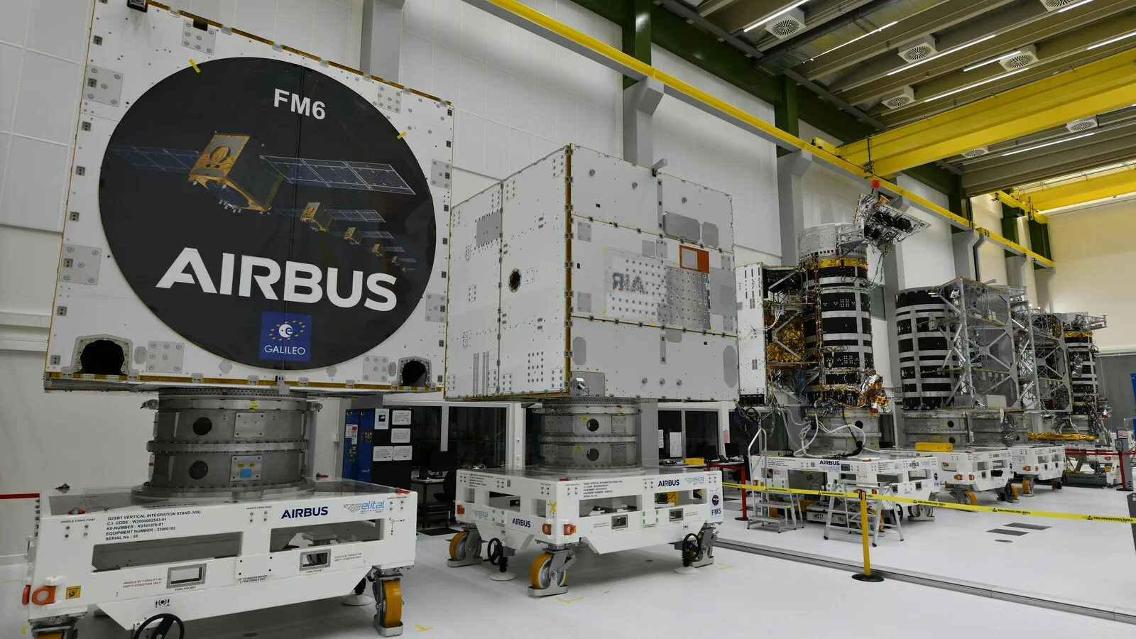 Pavilhão industrial branco com vários satélites Galileo de segunda geração em fila, em suportes com rodas, em diferentes fases de montagem