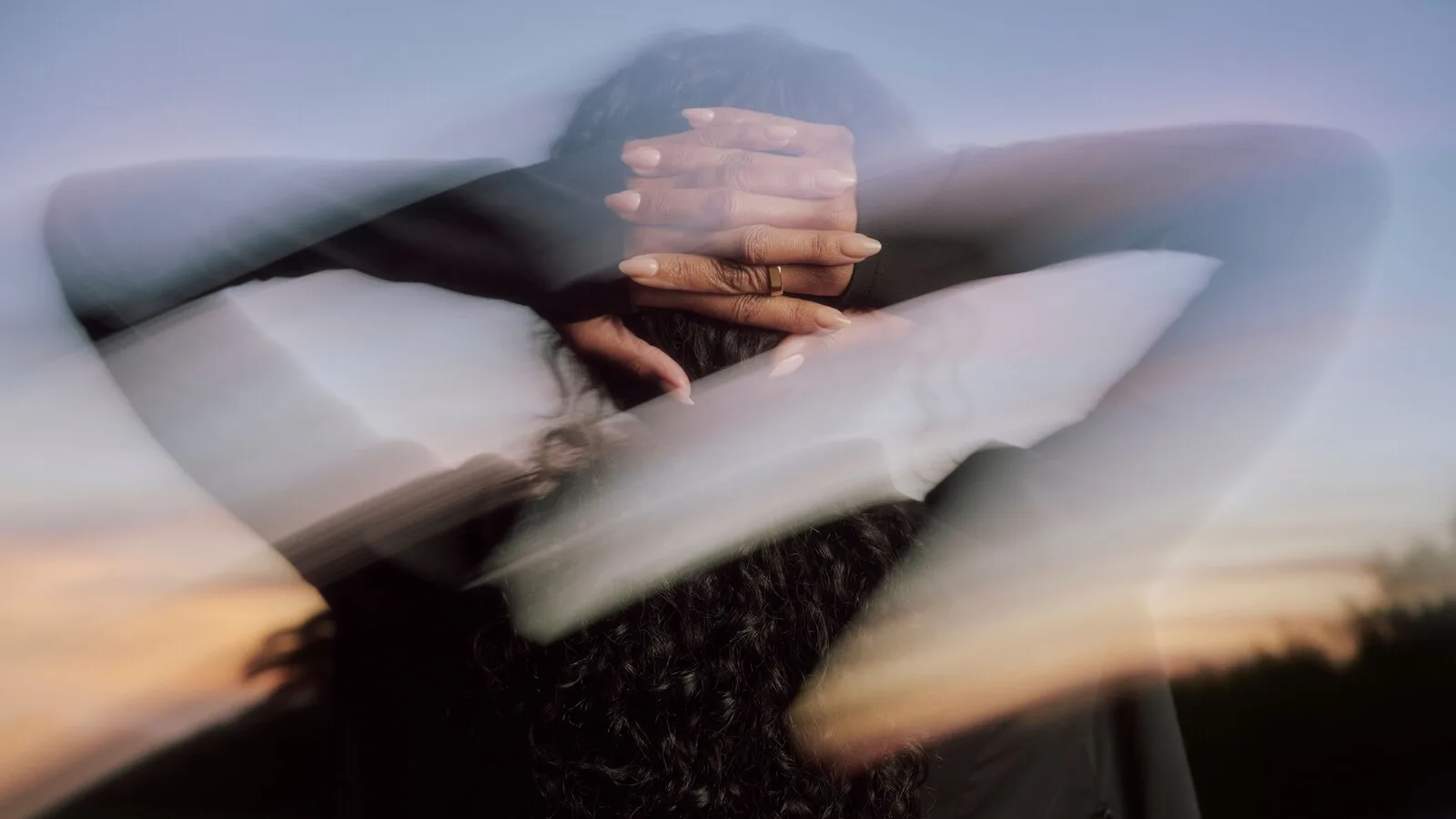 Mulher de costas com as mãos cruzadas atrás da cabeça e um anel Oura num dedo, numa fotografia arrastada com o céu do pôr do sol ao fundo
