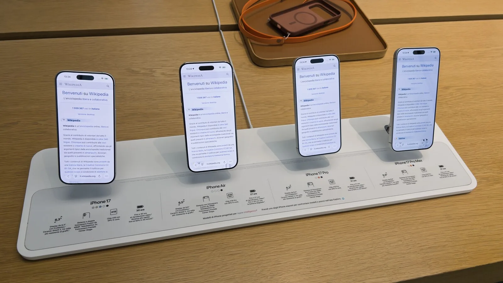 Quatro iPhone de pé num expositor branco de uma loja da Apple, todos com a página inicial da Wikipédia aberta no ecrã, sobre uma mesa de madeira