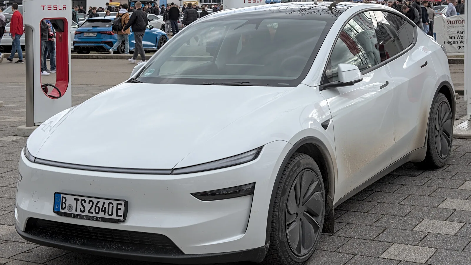 Tesla Model Y branco estacionado em frente a um posto Supercharger vermelho e branco, com pessoas e outros carros ao fundo