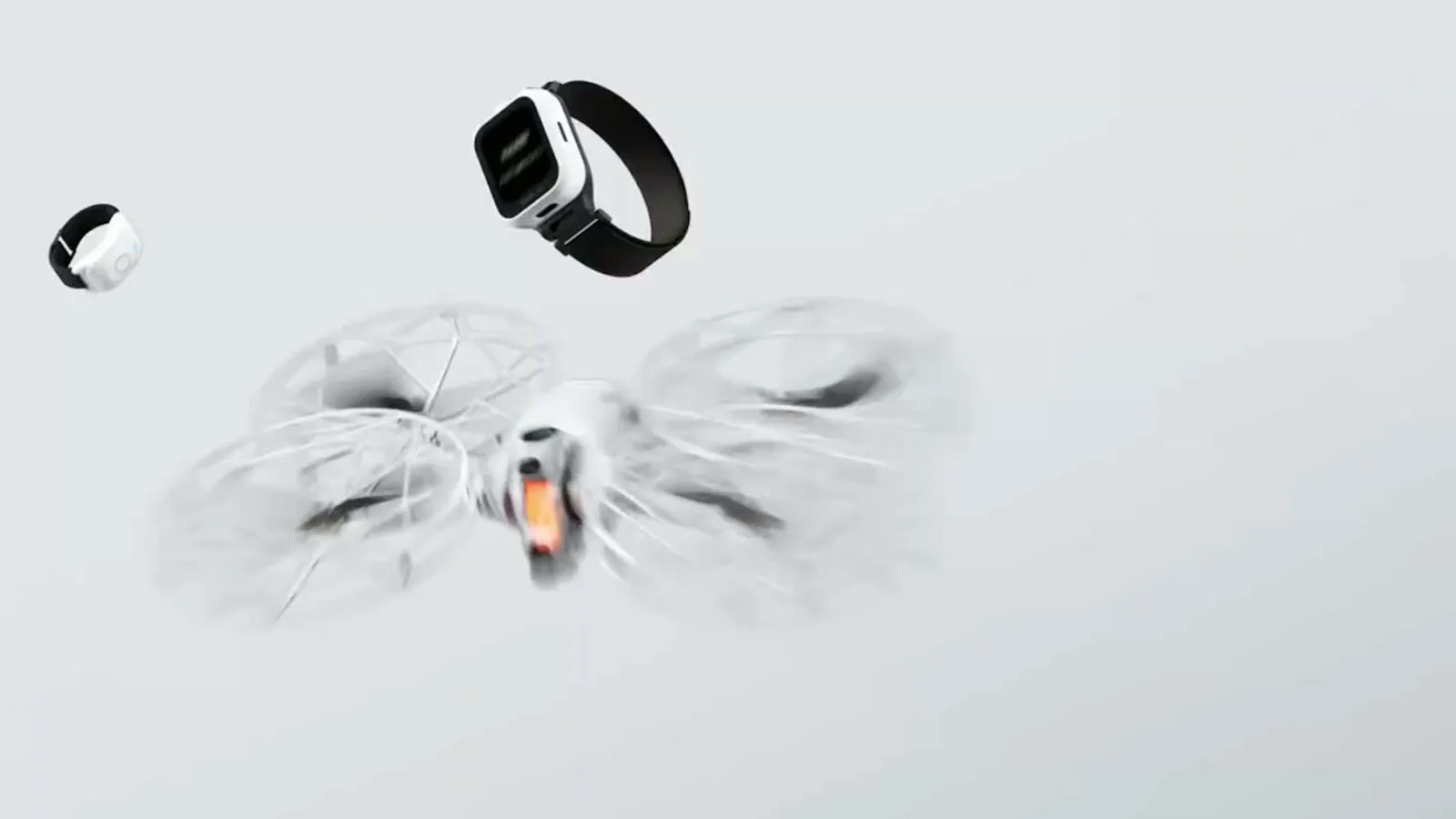 Drone branco com protetores circulares nas hélices, acompanhado por um relógio remoto com bracelete preta e um pequeno anel de controlo, sobre fundo cinzento claro