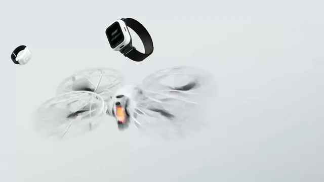 Drone branco com protetores circulares nas hélices, acompanhado por um relógio remoto com bracelete preta e um pequeno anel de controlo, sobre fundo cinzento claro