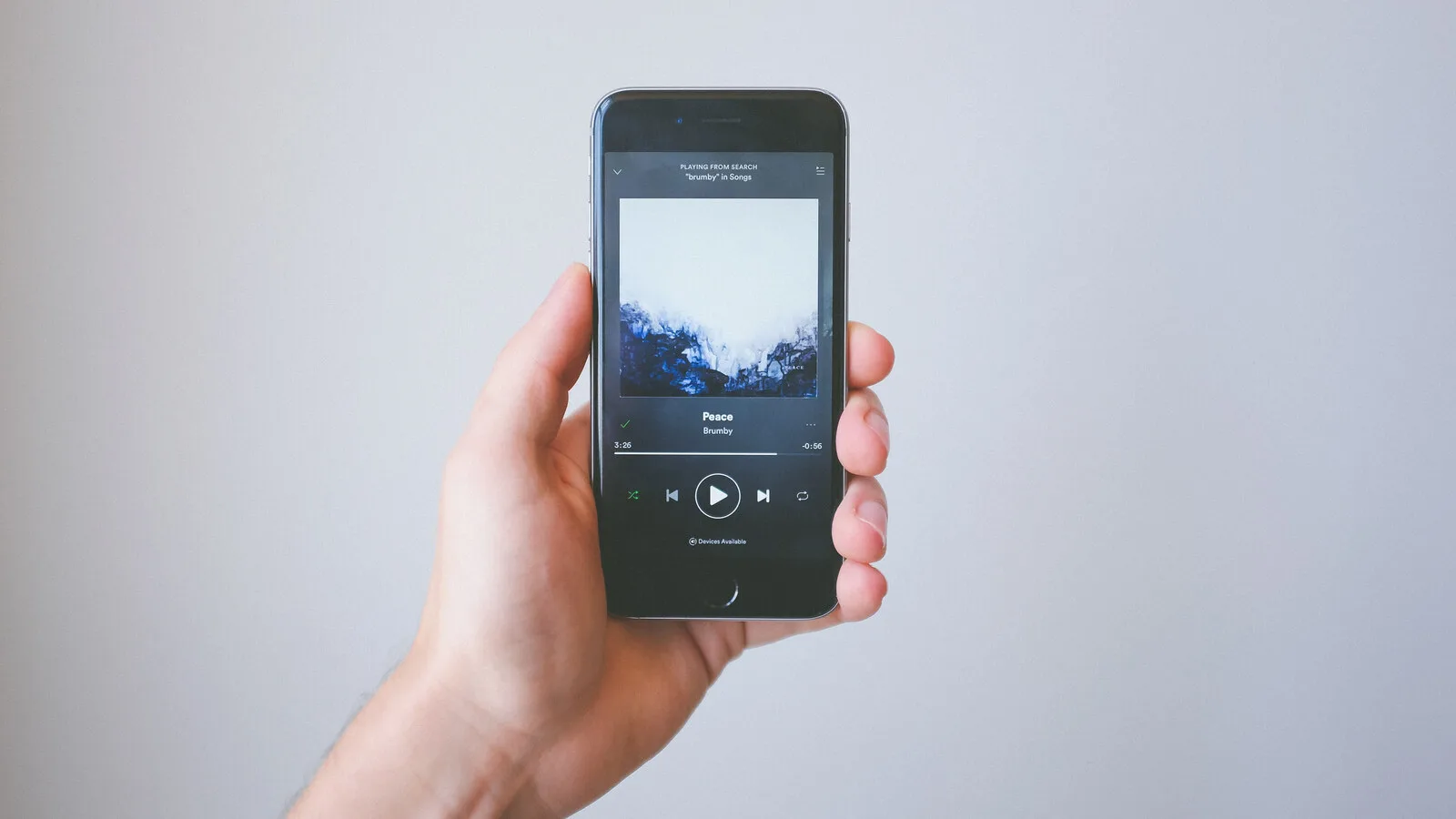 Mão a segurar um iPhone com o leitor do Spotify aberto, a mostrar a capa de um álbum e os botões de reprodução, sobre fundo cinzento